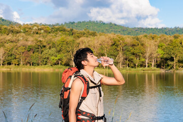 An asian man with backpacker smile and feeling fresh by drinking a bottle of water beside a lake in mountain alone