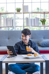 A handsome asian man is working from home using tablet in the living room in his home with snack and orange juice