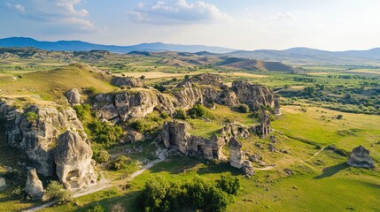 Aerial view of ancient stone formations surrounded by lush greenery and mountains under a clear blue sky in a rural landscape.