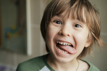 Cheerful funny toddler cute boy is showing tongue making faces at camera, fooling around, joking,...