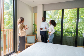 Obraz premium Real estate agent in business attire presents sunny bedroom to diverse young couple during property viewing consultation at tropical residential location.