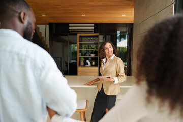 Real estate professional in beige blazer presents modern interior space to diverse couple during property viewing consultation at upscale residential location.