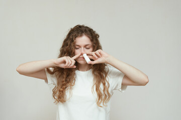 Fototapeta premium A young woman with curly hair in a white T-shirt uses a nasal spray, looking anxious and confused. Concept of health care, allergies, self-treatment, and battling cold or respiratory symptoms.