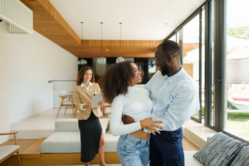 Young diverse couple celebrates property purchase in contemporary home interior while professional realtor observes their milestone achievement in casual business attire.