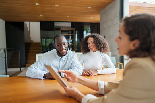 Professional real estate agent in business attire discusses property details with diverse couple at modern office. Digital tablet enhances interactive consultation in contemporary workspace.