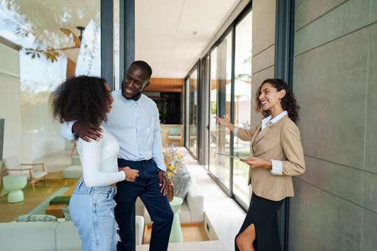 Real estate professional in business attire presents luxury property features to diverse couple during residential home tour in modern glass-walled space.
