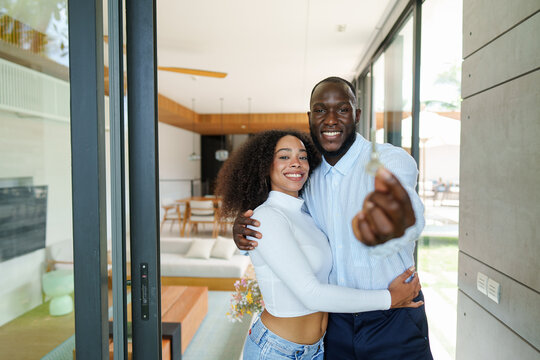 Diverse couple in casual attire celebrates milestone moment displaying house keys at their newly purchased residential property entrance.
