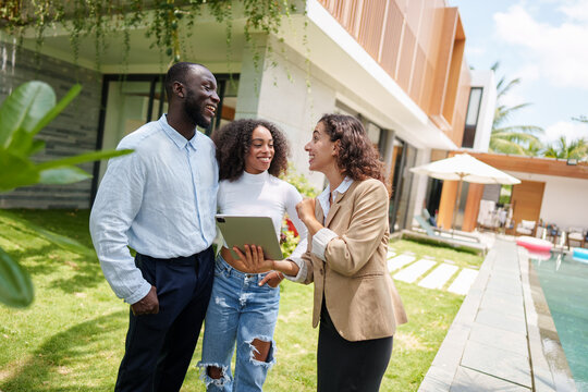 Real estate professional in blazer discusses property listings on tablet with diverse couple during outdoor consultation at modern residential complex.