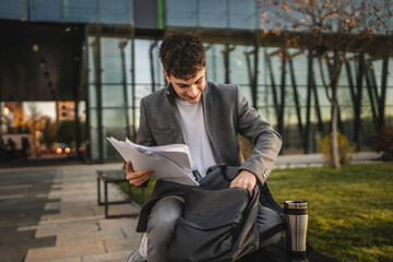 Fototapeta premium young man sit on bench with backpack and find documents and notes