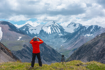 Man in vivid red jacket admire alpine scenery on sunlit green grassy hill near precipice edge. Guy on mountain pass enjoying view to few big snowy pointy peaks. Three large snow peaked tops far away.