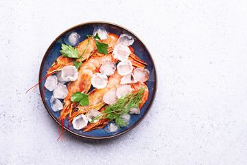Tiger shrimps with ice cubes, fresh dill twigs and parsley in dark blue bowl on light marble background top view.