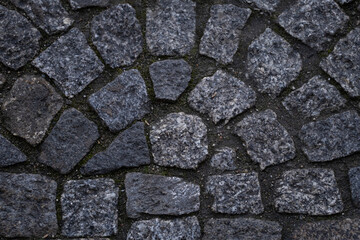 A textured close-up of gray cobblestones arranged in an uneven pattern. The natural stone surface showcases various shapes and tones, with green moss growing between the gaps.