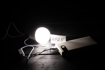 An energy-saving light bulb connected to an external battery and a card with the year 2025 written...