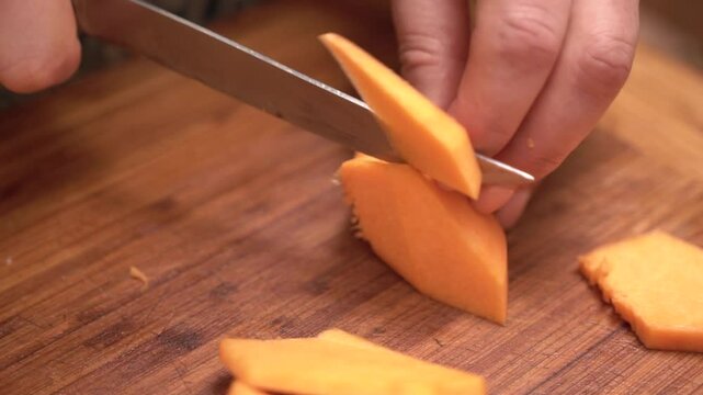 A person is cutting a piece of orange squash on a wooden cutting board. The squash is cut into slices, and the person is using a knife to cut it.