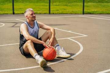 Fototapeta premium Young man sitting on basketball court, resting after playing, enjoying the outdoors on a sunny day