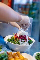 Male chef's hands with disposable gloves preparing a salad with grated radish, carrot, purple cabbage and fresh arugula and lettuce leaves on steel kitchen counter in a fish restaurant kitchen