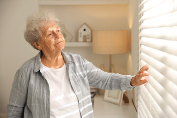 Fototapeta premium Loneliness concept. Sad senior woman looking through window blinds at home