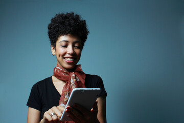 Cheerful afro american woman reading an eBook on a tablet. Short hair female model in front blue background with copy space. Suitable for literature, study, web search, online activity advertisements.