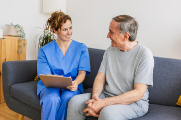Young female general practitioner in blue uniform talking with aged senior patient during checkup at home.