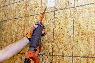 Craftsman using power cutting tool to make precise cuts in an oriented wooden plywood board wall for insulation installation.