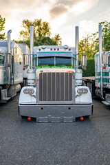 American idol white big rig classic semi truck tractor with vertical exhaust pipes standing in row with another semi trucks on truck stop parking lot at twilight time