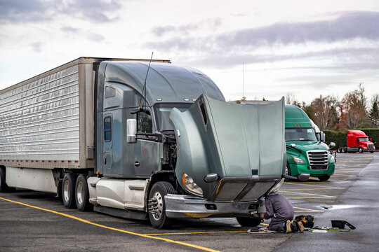 Truck driver repairing broken gray big rig semi truck with open hood and refrigerator semi trailer directly at the truck stop parking lot