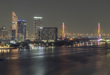 Fototapeta premium Nightscape city view with bright glowing lighting of Skyscrapers and Bhumibol bridge with Chao phraya river in the foreground. City scenery night of Bangkok in the sundown, use it as wallpaper, Poster