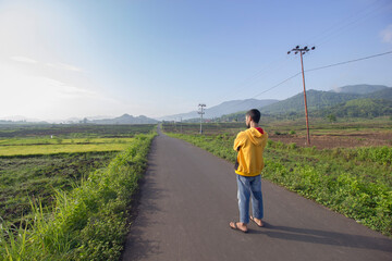 Traveler Walks Down Rural Road Through Green Rice Paddies towards Distant Mountains in Morning Sunlight