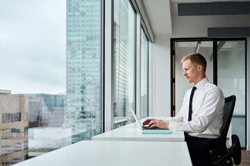 Young business man executive wearing tie working on laptop in skyscraper office at desk near big...