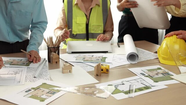 Smart architect and safety engineer discussion about building blueprint on table with architectural document, measurement tools and wooden block scatter around at modern office. Closeup. Delineation.