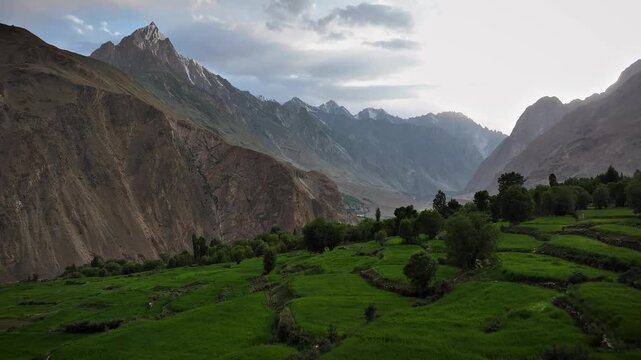 Aerial footage in Pakistan panning over lush green agriculture towards a winding river and the rugged Karakoram Mountains.