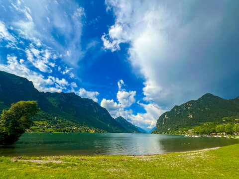 Stunning landscape featuring a calm lake reflecting the cloudy sky. Majestic mountains rise in the distance, framed by lush greenery, creating a tranquil and picturesque scene. Lake Idro, Crone, Italy