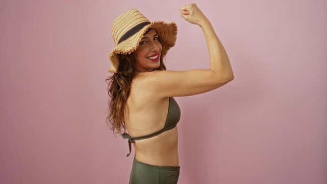 Middle-aged woman flexing muscles wearing a bikini and straw hat against a pink isolated background