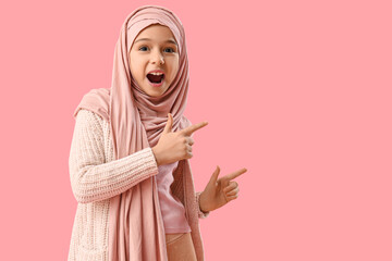 Shocked little Muslim girl in hijab pointing at something on pink background