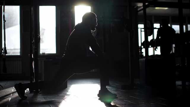 Professional athlete stretching legs dynamically, muscular silhouette backlit by warm morning sunlight streaming through expansive gym windows