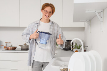 Young man with water filter jug showing thumb-up near sink in kitchen