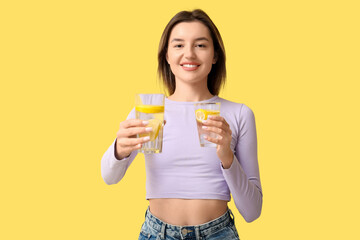 Beautiful young woman with glasses of lemon infused water on yellow background