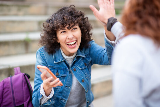 Portrait of a young woman giving high-five to a friend while holding phone outdoors