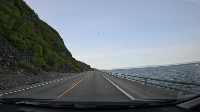 Driving through the tundra on E69 in northern Norway. Rocky hills with sparse trees and grass rise on the left, while the sea stretches along the right, showcasing raw and rugged nature.