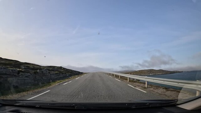 Driving along Norway's E69 road in the north. The sea is on the right, with low clouds and mist above. Surrounded by rugged landscapes, rocks, grass, moss, lichens, and small lakes.