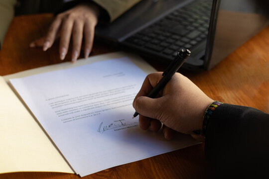 Close-up of a businessman's hand signing a document or contract at an office desk with a laptop of a woman with whom he is doing business and closing deals