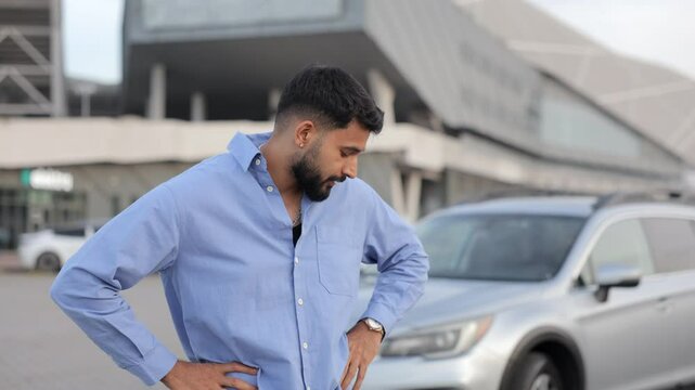 Bearded upset driver standing in front of broken car after road accident in city. Tired, disappointed guy rubbing forehead, shaking head, thinking about car crash. Concept of car break down, emotions.