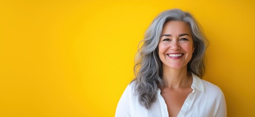 Smiling woman with gray hair against vibrant yellow background, showcasing confidence and beauty in aging, wearing white shirt, natural light, copy space for text
