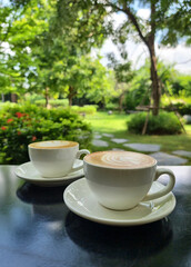 Vertical shot of the two cups of hot latte art coffee on a wooden table and the green garden background.