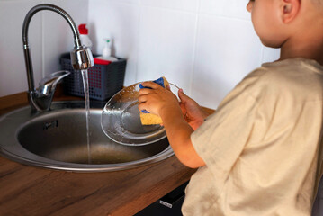 Funny 5-year-old boy is washing dishes standing at the sink. Housework assistant, the concept of work.