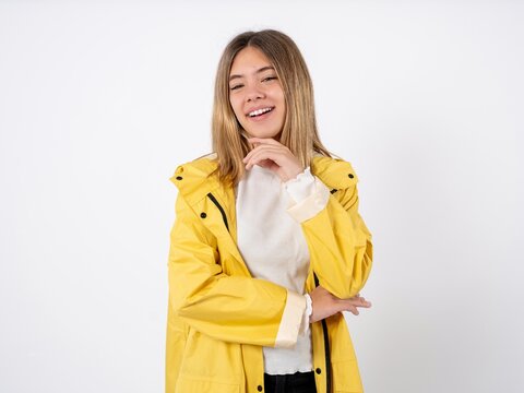 Optimistic Beautiful teen girl wearing rain coat keeps hands partly crossed and hand under chin, looks at camera with pleasure.