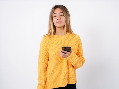 beautiful caucasian teen girl wearing orange sweater holds mobile phone in hands and rejoices positive news, uses modern cellular