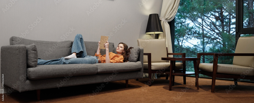 Wall mural Young woman relaxing on a cozy sofa, reading a book, with a modern interior design and large windows, evoking tranquility and comfort in a serene environment