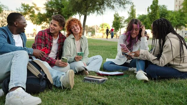 University student friends sitting on the grass on campus, laughing using mobile phone app sharing content on social media