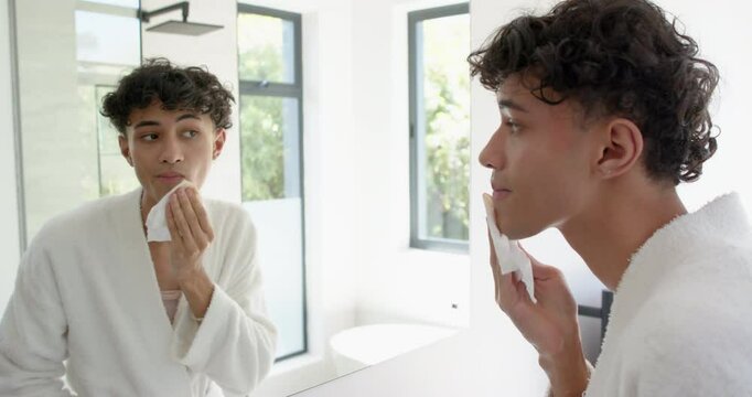 Looking in mirror, person in bathrobe wiping face with tissue in bathroom